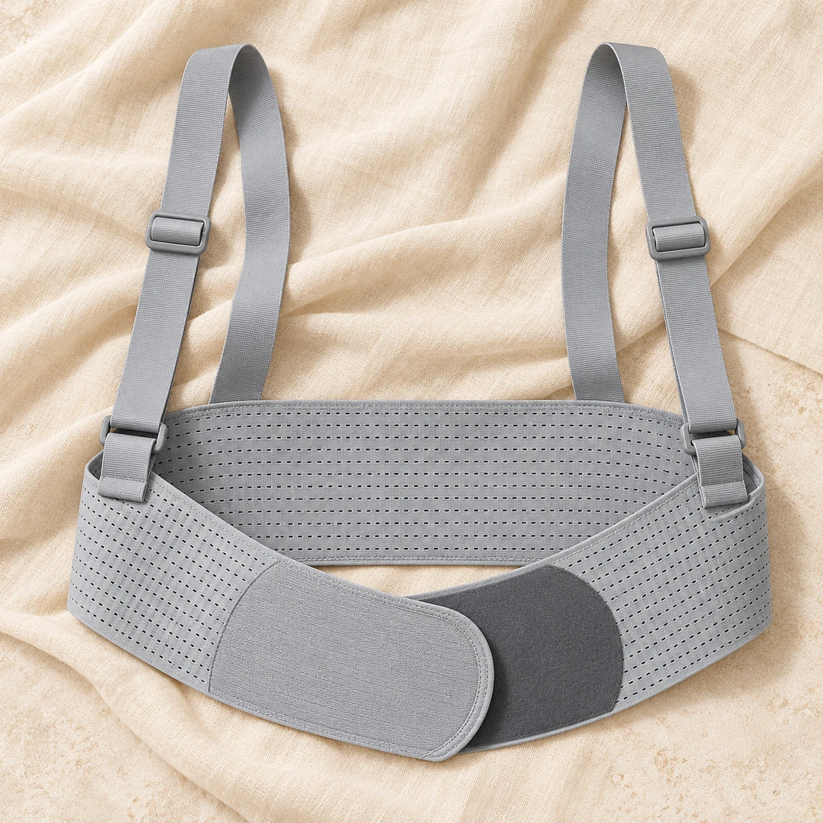 grey perforated under-bump support belt with wide back section, overlapping rounded fastening panels and two adjustable shoulder straps with matching rectangular sliders, product on cream linen